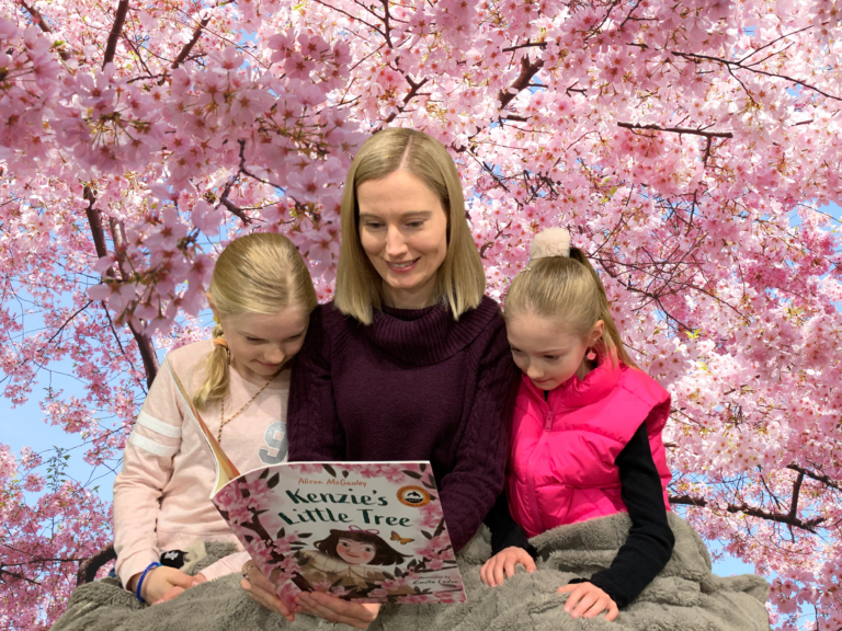 Alison McGauley and her two daughters read the book "Kenzie's Little Tree" together. A blooming tree is in the background.