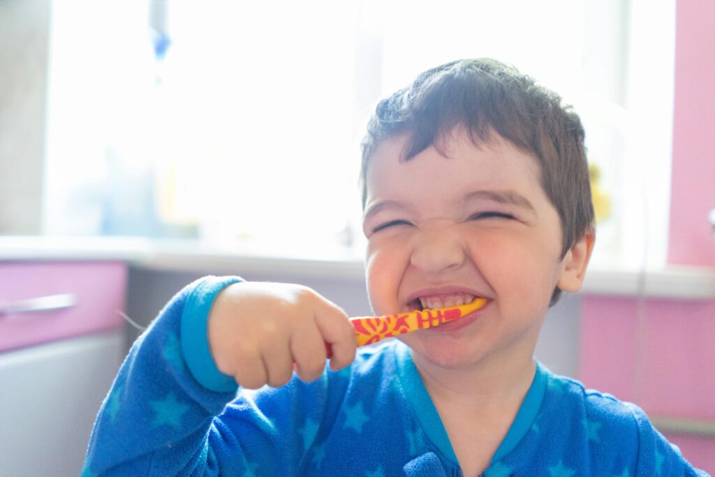 A little boy with brown hair in blue pajamas is squinting and smiling as he brushes his teeth with an orange toothbrush