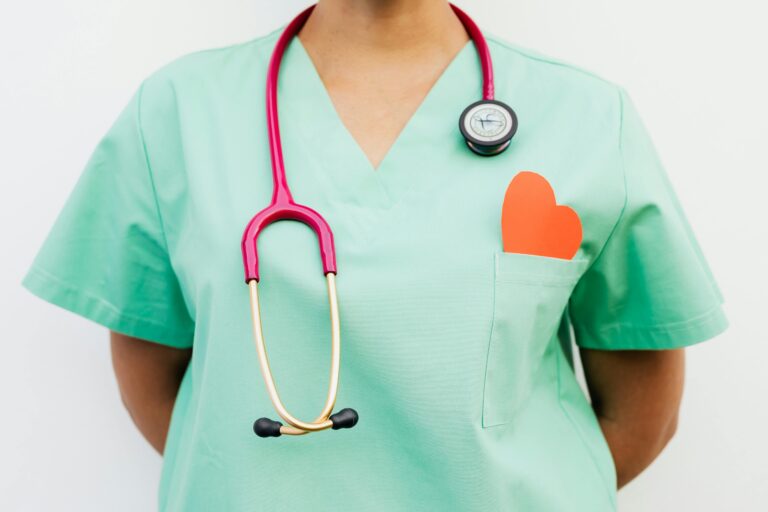 The torso of a woman in green scrubs with a pink stethoscope around her neck. There is a paper heart sticking out of her breast pocket.