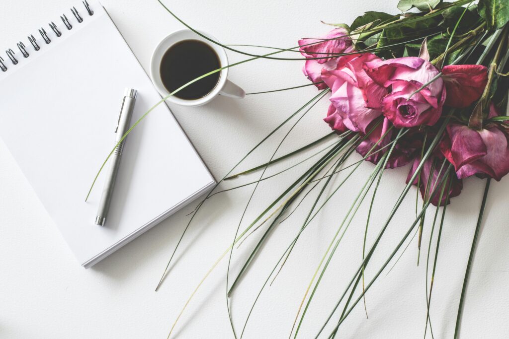 A birds-eye view of a blank notebook, a pen, a cup of coffee, and a bouquet of roses.